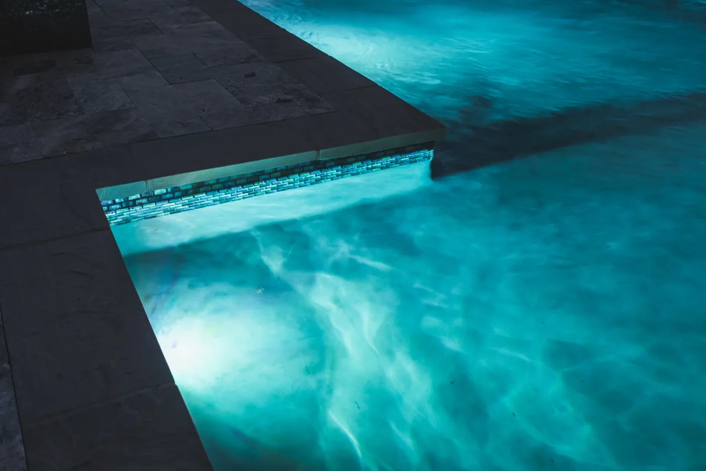 LED-lit pool edge with glass tile band and travertine coping at night — Tarkington Residence
